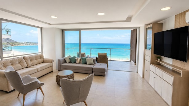 A modern living room with a large window overlooking a beach, a sofa set, a television, and a dining area. - Luxury Beachfront Apartment in Chipipe – Stunning Views & Top Amenities (Salinas)