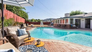 Ein Schwimmbecken mit Liegestühlen und Sonnenschirmen, umgeben von einer Ziegelterrasse und einem Gebäude mit großen Fenstern.