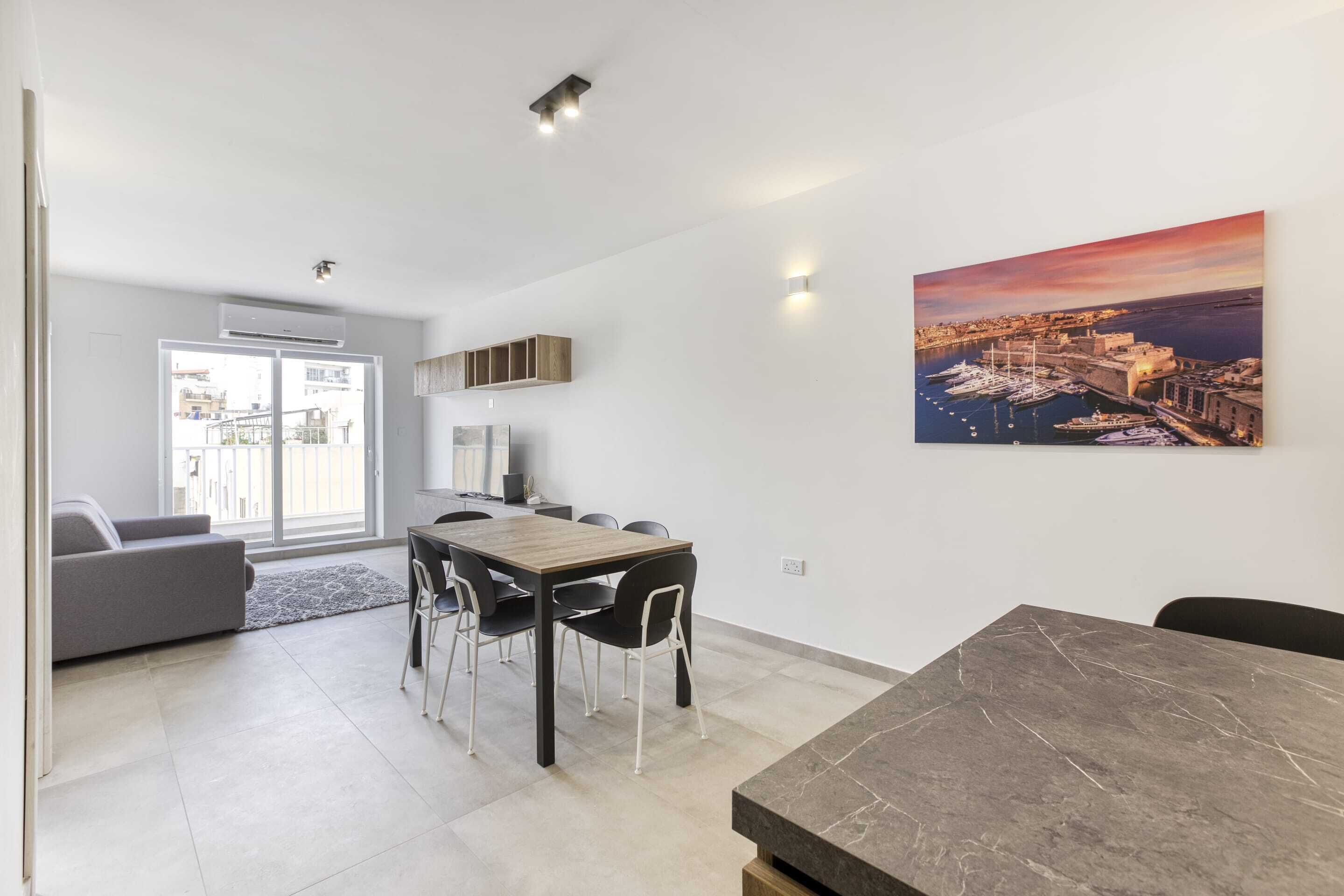 Uma sala de estar moderna com área de jantar, um sofá cinza, uma mesa de jantar de madeira e uma pintura grande de uma paisagem costeira.