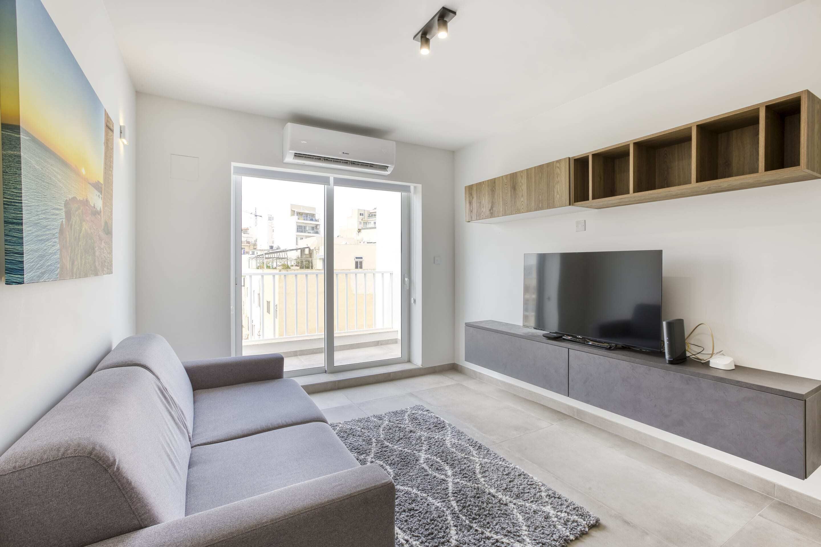 A modern living room with a grey sofa, a flat-screen TV on a wooden shelf, and a large window with a view of buildings.