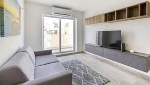 A modern living room with a grey sofa, a flat-screen TV on a wooden shelf, and a large window with a view of buildings. - Gzira Suite 14 (Gzira)