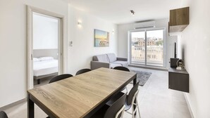 A modern living room with a wooden dining table, grey sofa, and a city view through the glass door. - Gzira Suite 14 (Gzira)