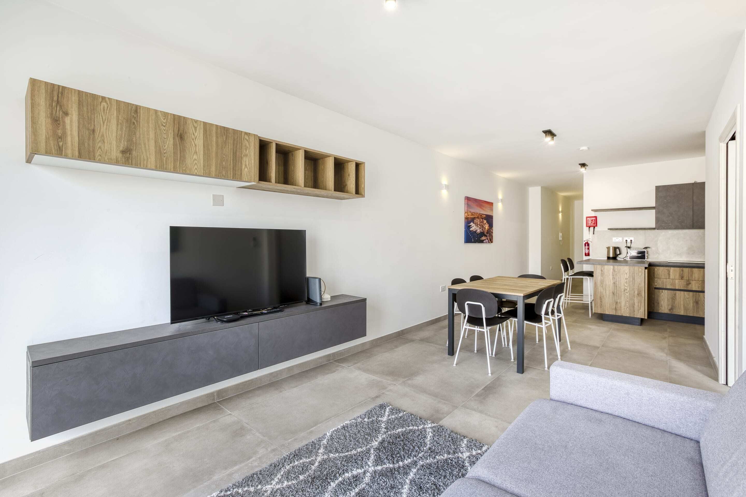 A modern living space with a flat-screen TV, built-in shelves, a dining area with chairs, and a kitchen area with cabinets and a stove.