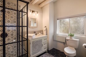 Un baño con cabina de ducha de vidrio, un lavabo blanco con un espejo decorativo, un inodoro con ventana y un suelo estampado.