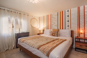 Un dormitorio con cama, mesita de noche, lámpara, pared con diseños geométricos y ventana con cortinas.
