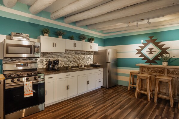 Una cocina con armarios blancos, una estufa de acero inoxidable, un microondas y un refrigerador. Hay taburetes de madera y una pared decorativa con patrones geométricos.