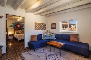 Un salón con un sofá azul, una mesa de centro de madera y una alfombra estampada. Hay obras de arte abstracto en las paredes y una ventana con vistas a un edificio.