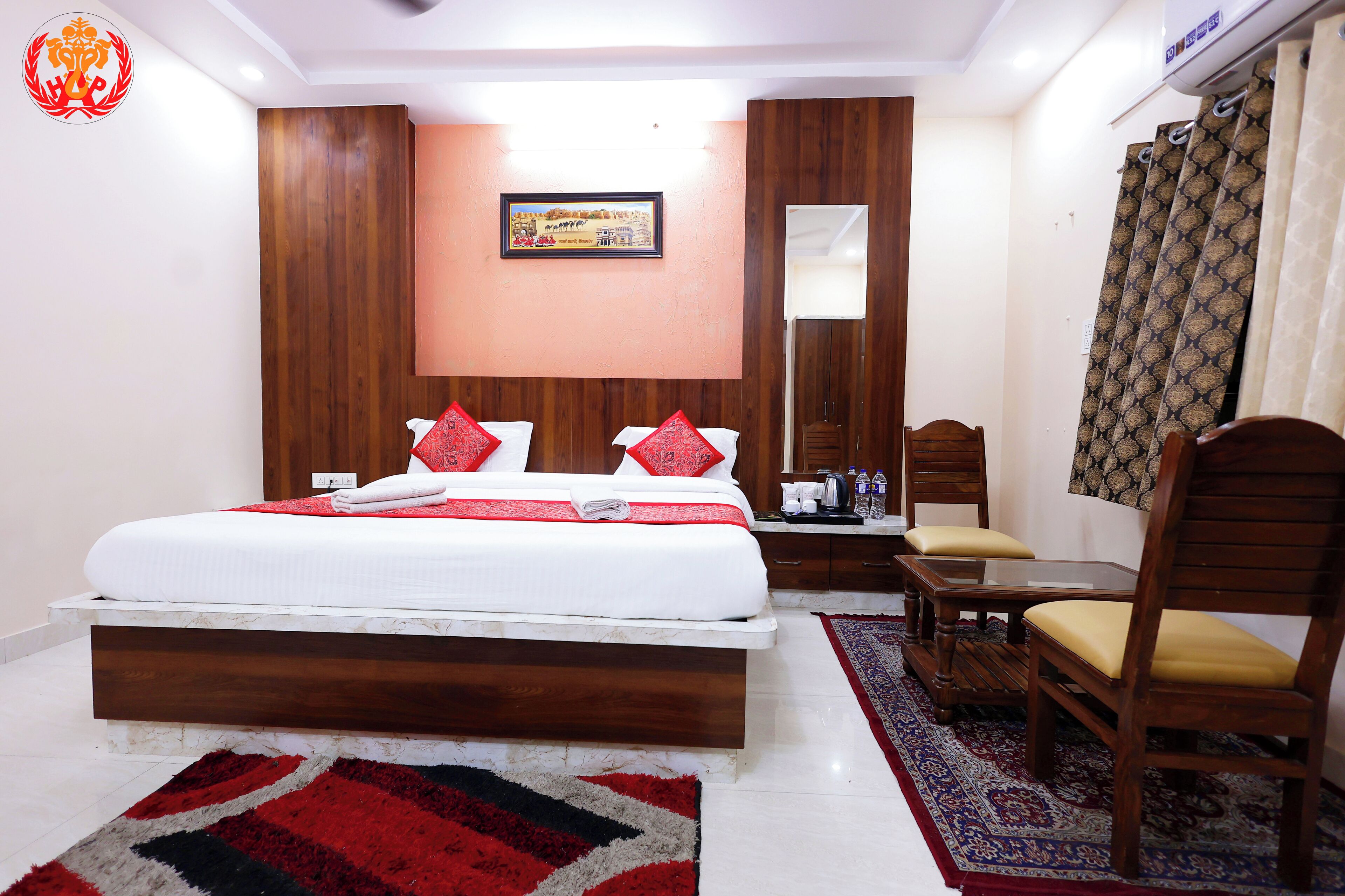 Ein Hotelzimmer mit einem großen Bett, einem hölzernen Kopfteil, einem kleinen Tisch mit Stühlen, einem Spiegel und einem gemusterten Teppich.