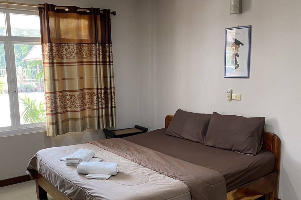 A neatly made bed with a wooden frame and a beige and brown bedspread, a bedside table with a lamp, and a window with curtains.