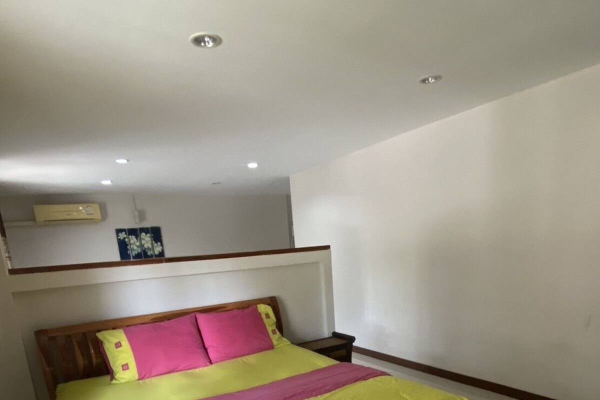 A neatly made bed with a bright yellow bedspread and pink pillows, a wooden headboard, and a small bedside table.