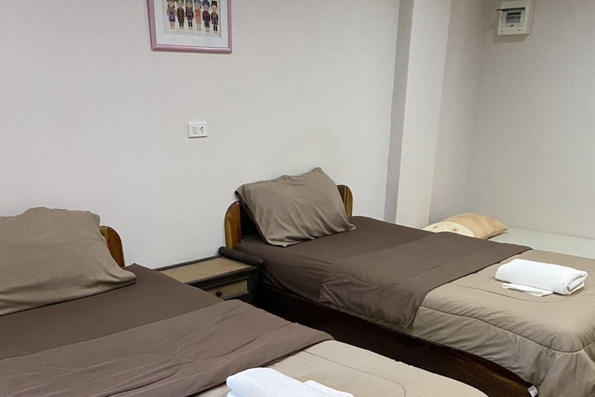 A hotel room with two single beds, each with a wooden frame and beige bedding. There is a framed picture on the wall above the beds, and a small electrical outlet is visible on the wall.