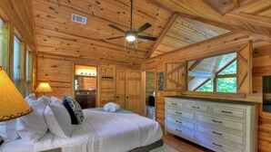 Une chambre dans une cabane en bois, avec un lit, une commode, un ventilateur de plafond et de grandes fenêtres.