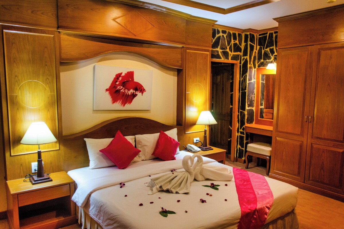 A hotel room with a wooden headboard, a bed with red and white pillows, a neatly made bed with a towel and flowers, a nightstand with a lamp, and a wooden wardrobe.