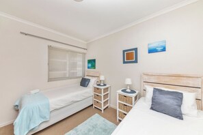A bedroom with two beds, a window with blinds, a bedside table with a lamp, and a wall-mounted picture.