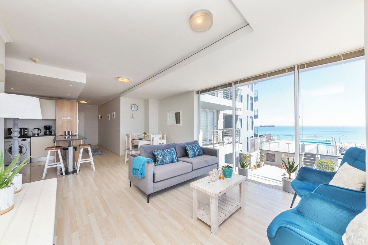 A modern living space with a sofa, coffee table, and chairs, a kitchen area, and a balcony with a view of the sea.