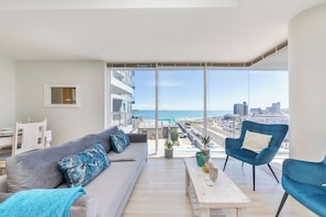 A balcony with a sofa, chairs, and a table, overlooking a cityscape and the sea.