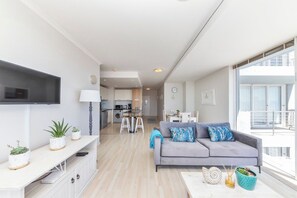 A modern living room with a grey sofa, a flat-screen TV mounted on the wall, and a balcony with a railing.
