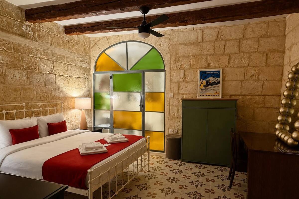 Quarto com cama, um armário verde, espelho decorativo e porta com painéis de vidro coloridos.