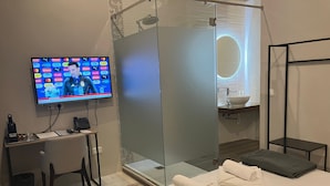 A hotel room with a TV displaying a news channel, a glass-enclosed shower, a bed with towels, a desk with a phone, and a small table with a trash bin. - Mulberry Suites by The LVH Collection  (Valletta)