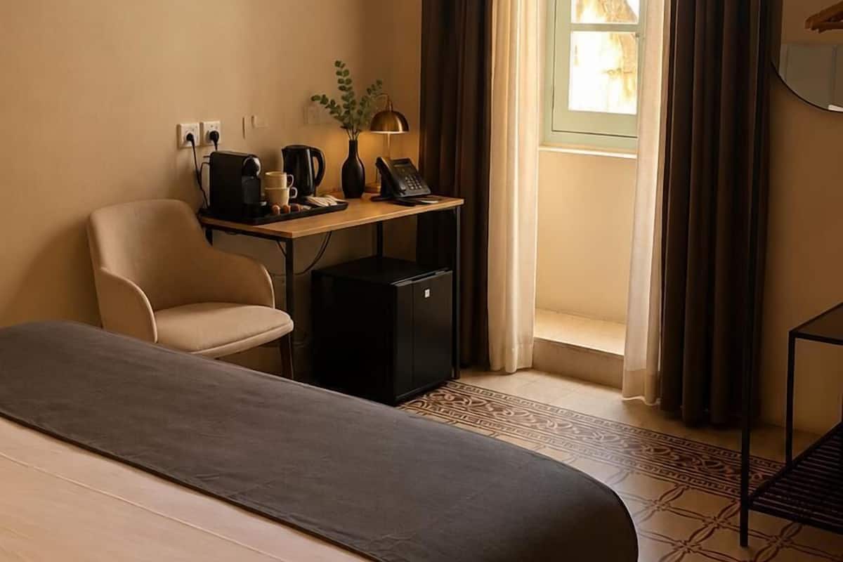 Quarto de hotel com cama, escrivaninha com computador, cadeira e janela com cortinas.