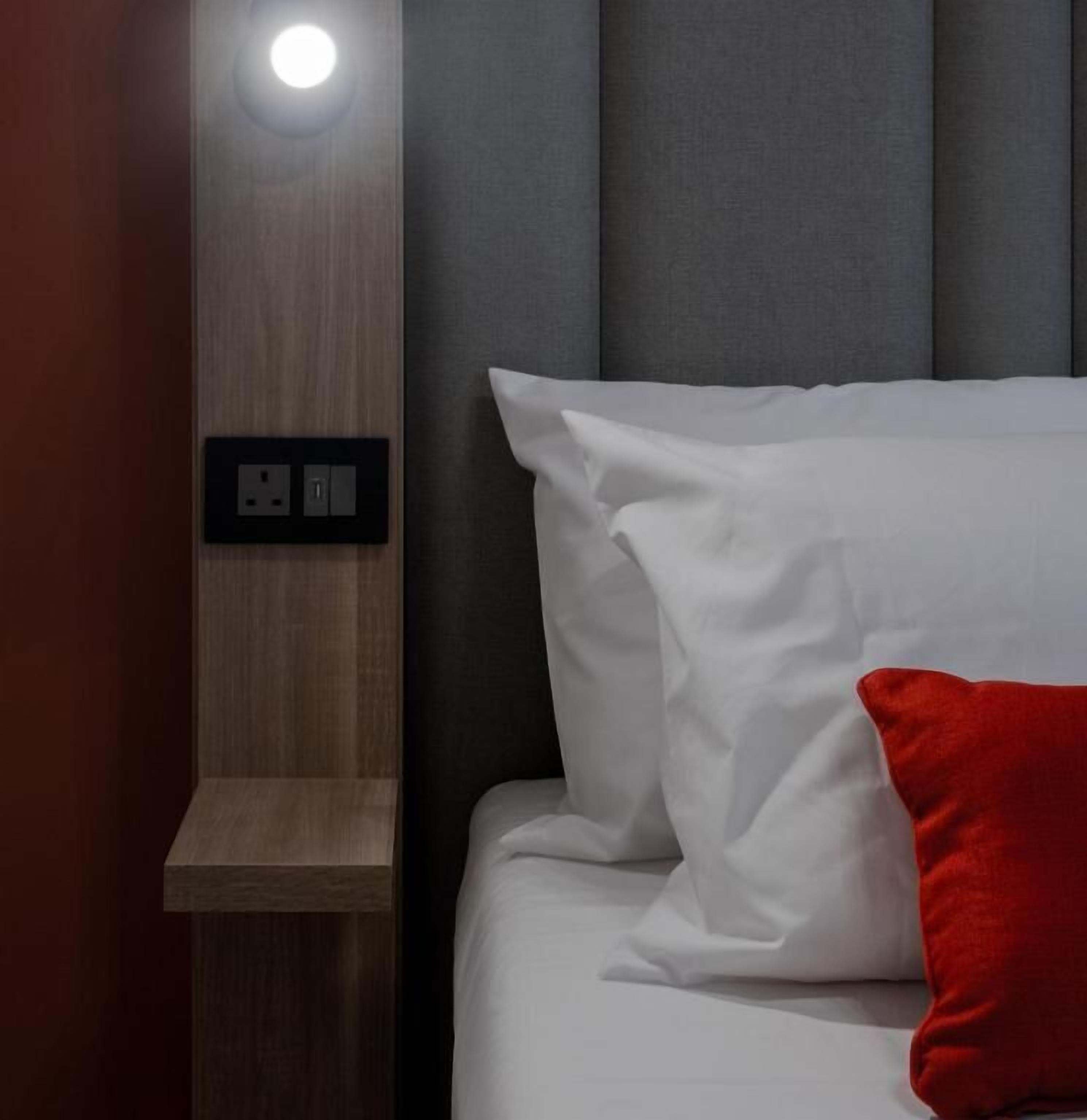 Una cama bien hecha con ropa de cama blanca y una almohada roja, una mesita de noche de madera con una lámpara y un enchufe en la pared.