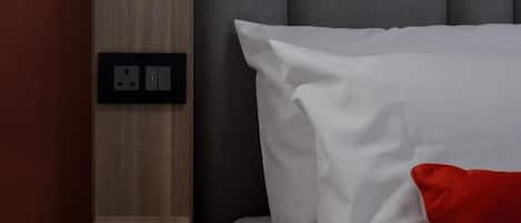 Una cama bien hecha con ropa de cama blanca y una almohada roja, una mesita de noche de madera con una lámpara y un enchufe en la pared.