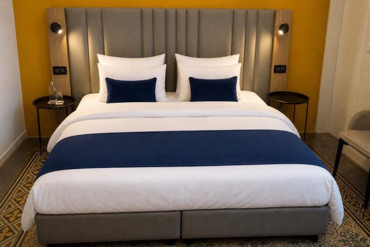 Uma cama grande com cabeceira cinzenta, roupa de cama branca e azul, e duas mesas de cabeceira com abajures.