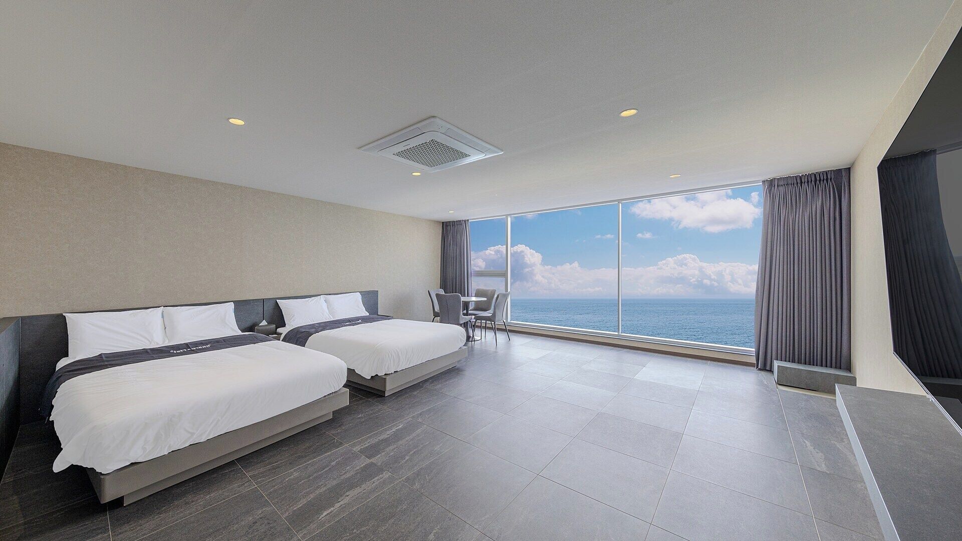 A modern hotel room with two beds, a small table, and a view of the sea.