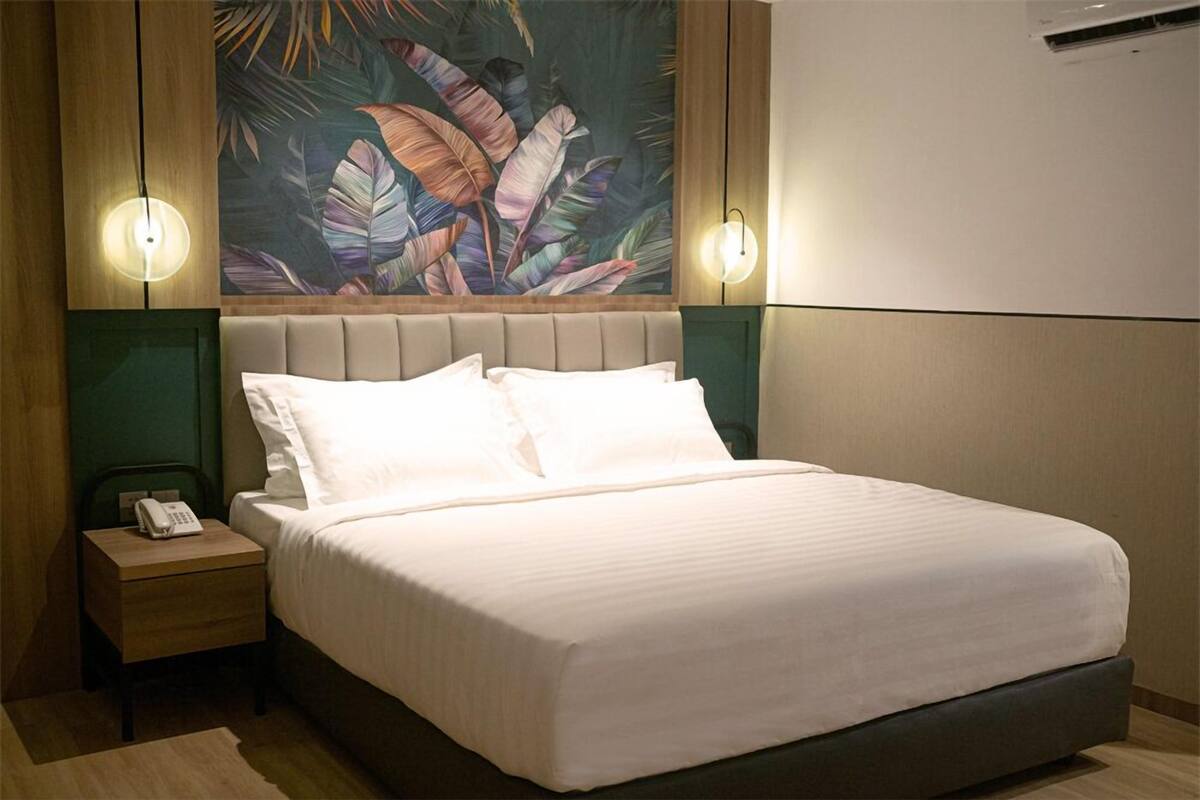 Una cama bien tendida con sábanas blancas en una habitación con un mural de hojas tropicales en la pared y dos lámparas fijadas a la pared.