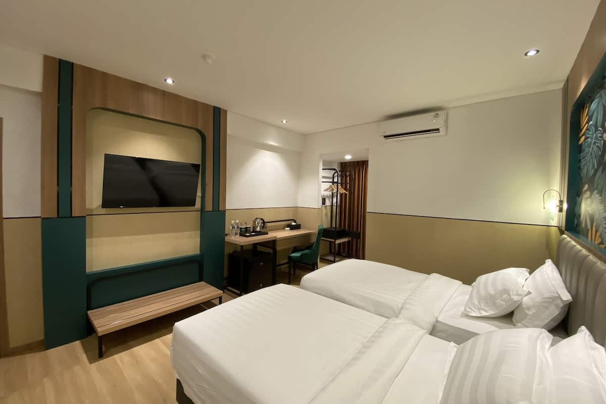 Habitación de hotel con dos camas, un escritorio con silla, un banco, un televisor y un equipo de aire acondicionado.