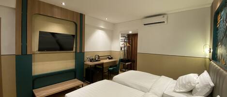 A hotel room with two beds, a desk with a chair, a bench, a TV, and an air conditioning unit.