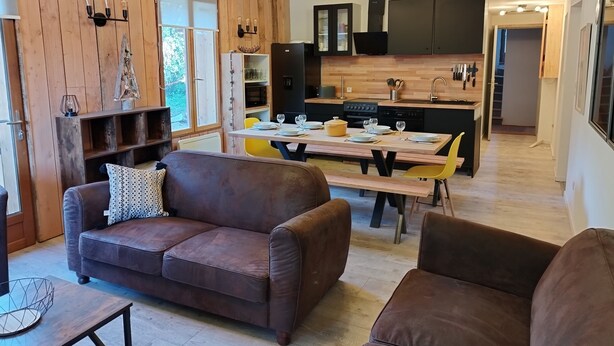 A modern living room with a brown leather sofa, a wooden dining table set for four, and a kitchen in the background with black cabinets and a wooden countertop.