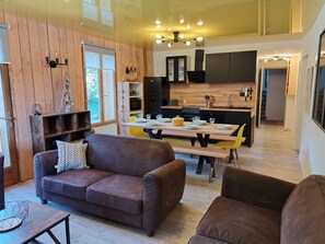 Uma sala de estar moderna com um sofá de couro marrom, uma mesa de jantar de madeira para quatro pessoas e uma cozinha ao fundo com armários pretos e bancada de madeira.
