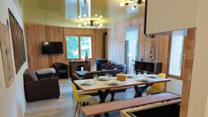 A cozy dining area with wooden walls and a large window, a television mounted on the wall, and a dining table set with plates and glasses.