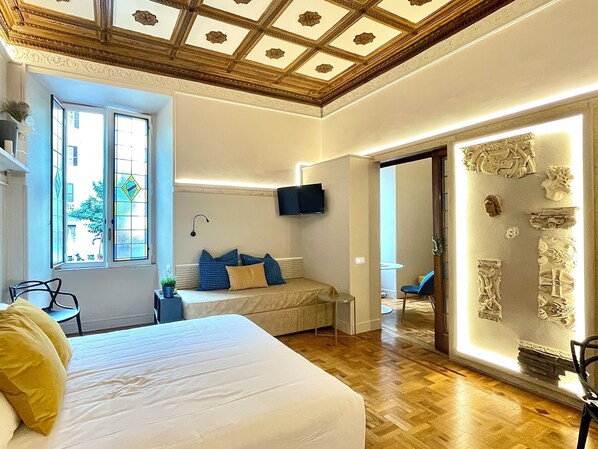 A spacious bedroom with a large bed, a TV mounted on the wall, and a window with a view of the outside.