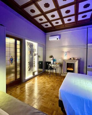 A bedroom with a wooden floor, a bed, a desk with a chair, a fireplace, and a large mirror.