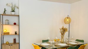 Un coin repas avec une table en verre dressée pour six personnes, entourée de chaises colorées. Un meuble en bois avec des étagères est visible sur la gauche, et un lampadaire est fixé au mur au-dessus de la table.