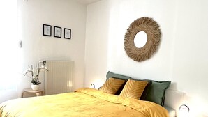 Une chambre avec un grand lit, une table de chevet avec une lampe, un objet décoratif fixé au mur et une petite plante posée sur une table d’appoint.