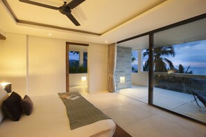 Une chambre moderne avec un grand lit, un ventilateur de plafond et un balcon offrant une vue sur les palmiers et l’océan.