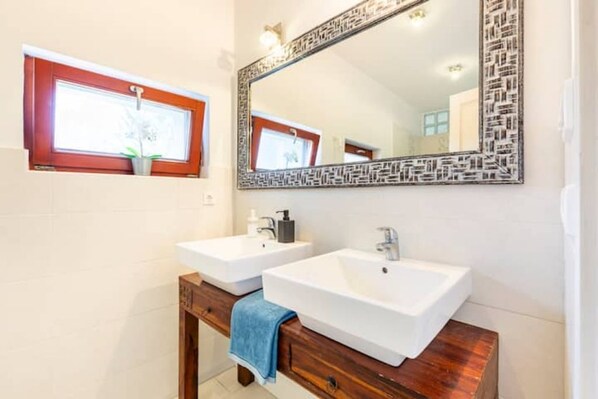 A bathroom with two white sinks, a wooden vanity, a large mirror with a decorative frame, and a window with a red frame.