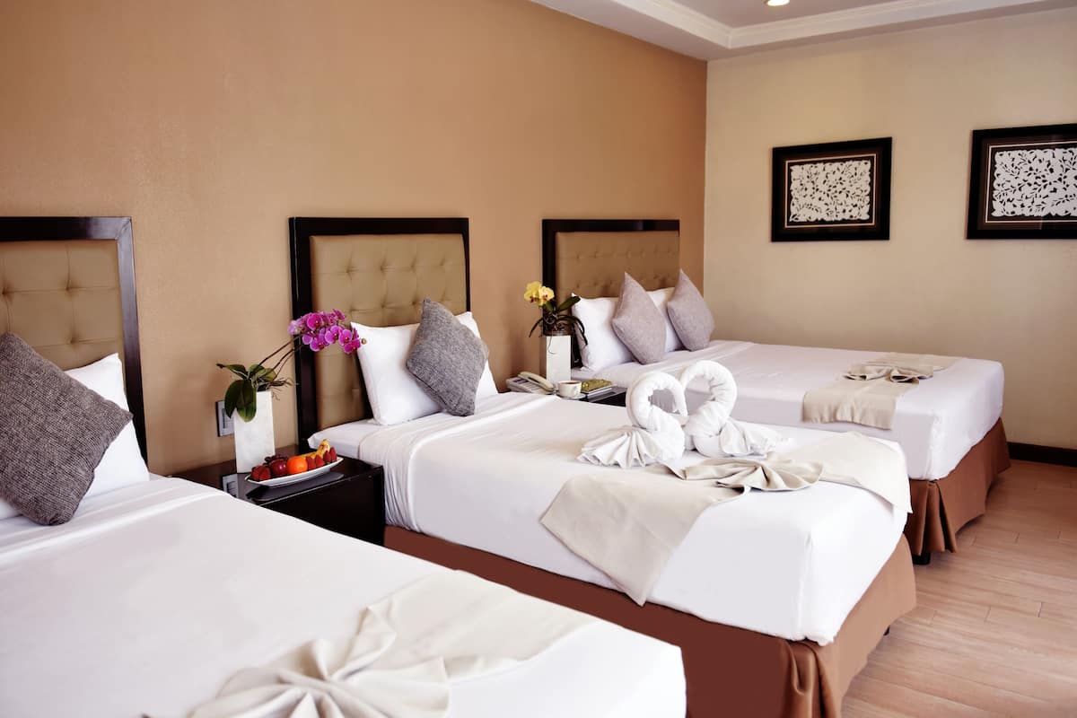 A hotel room with two beds, each with a headboard, white bedding, and decorative towels. There is a nightstand with a vase of flowers and a plate of fruit.