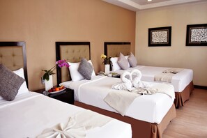 A hotel room with two beds, each with a headboard, white bedding, and decorative towels. There is a nightstand with a vase of flowers and a plate of fruit.