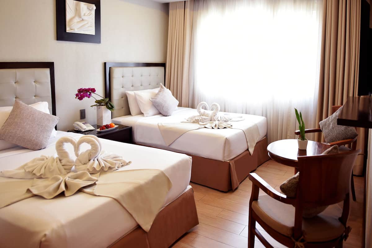 A hotel room with two beds, each with a neatly made bedspread and pillows. There is a small table with a vase of flowers and a lamp.