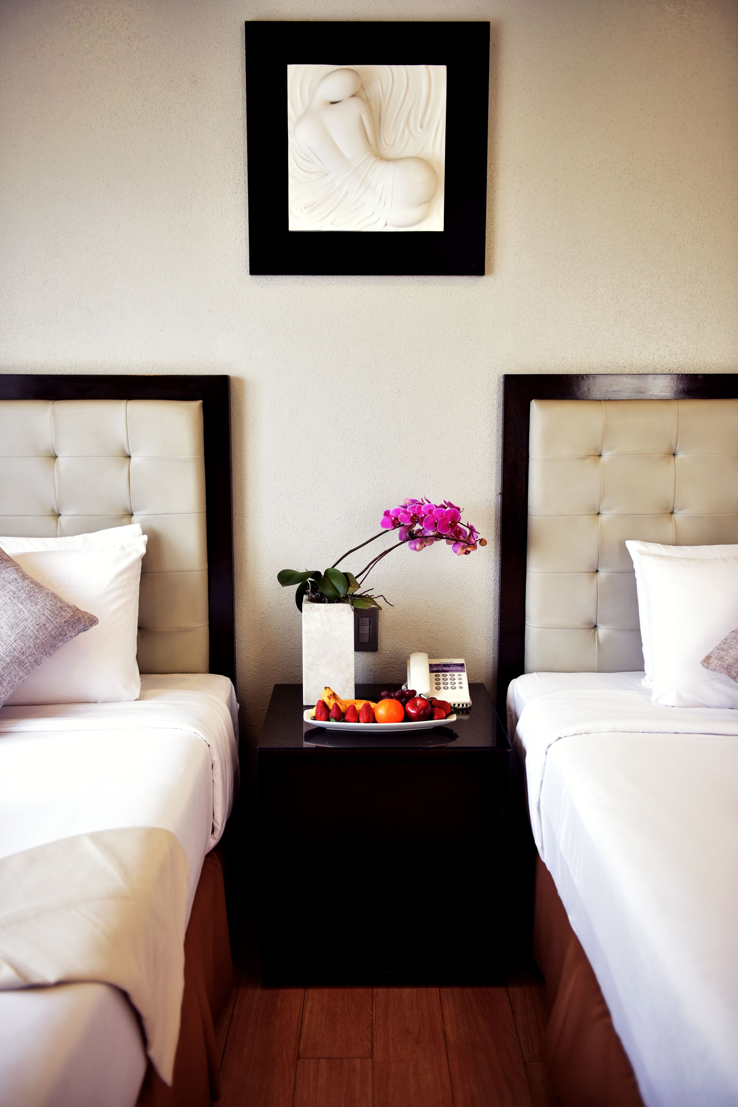 A hotel room with two beds, a nightstand with a vase of flowers, and a framed artwork on the wall.