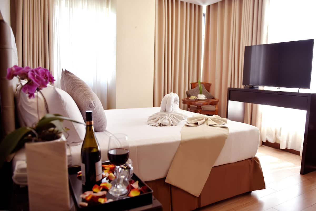 A hotel room with a bed, a TV, a tray with fruit and wine, and a vase with flowers.