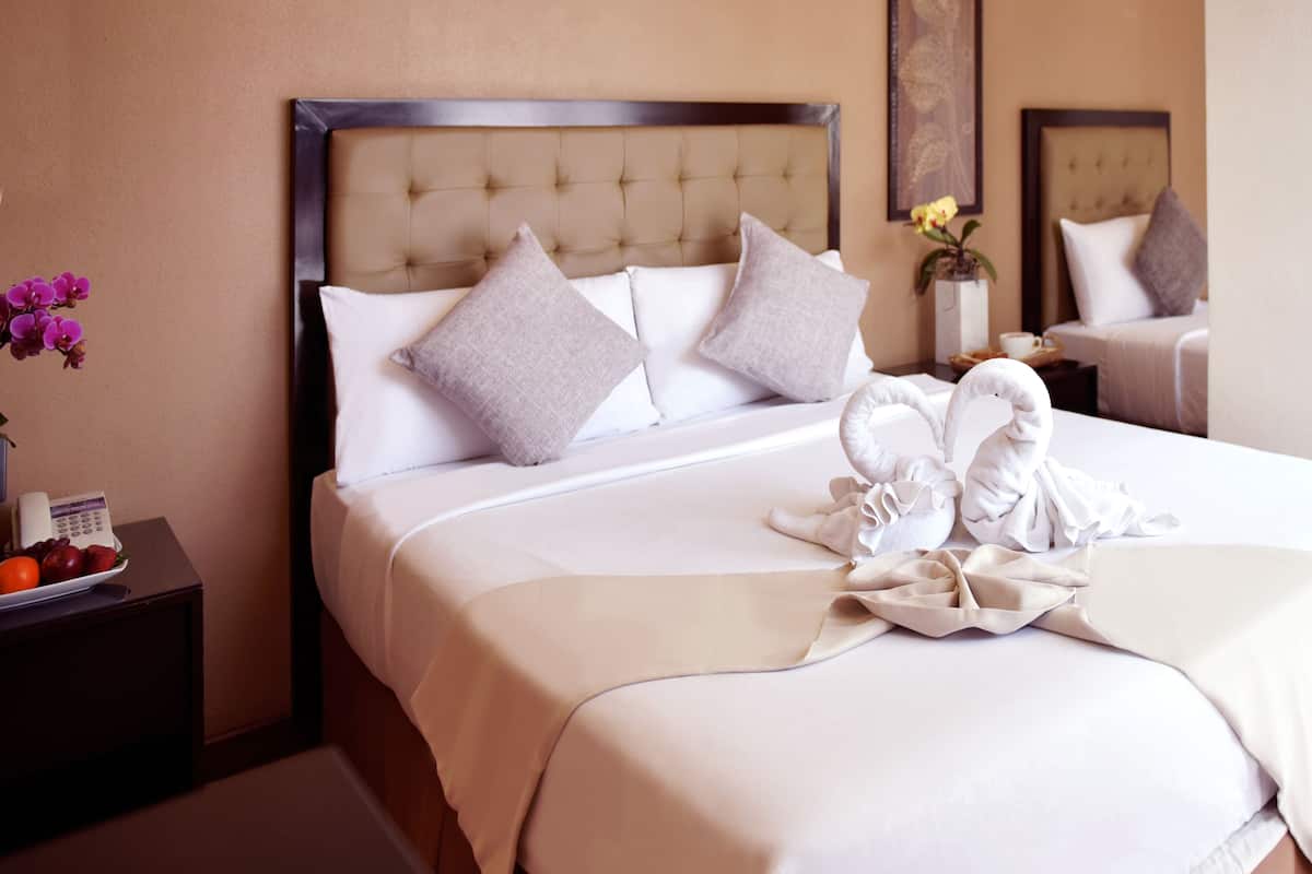 A neatly made bed with a tufted headboard, white linens, and two pillows. A small table beside the bed holds a vase with flowers and a plate of fruit.