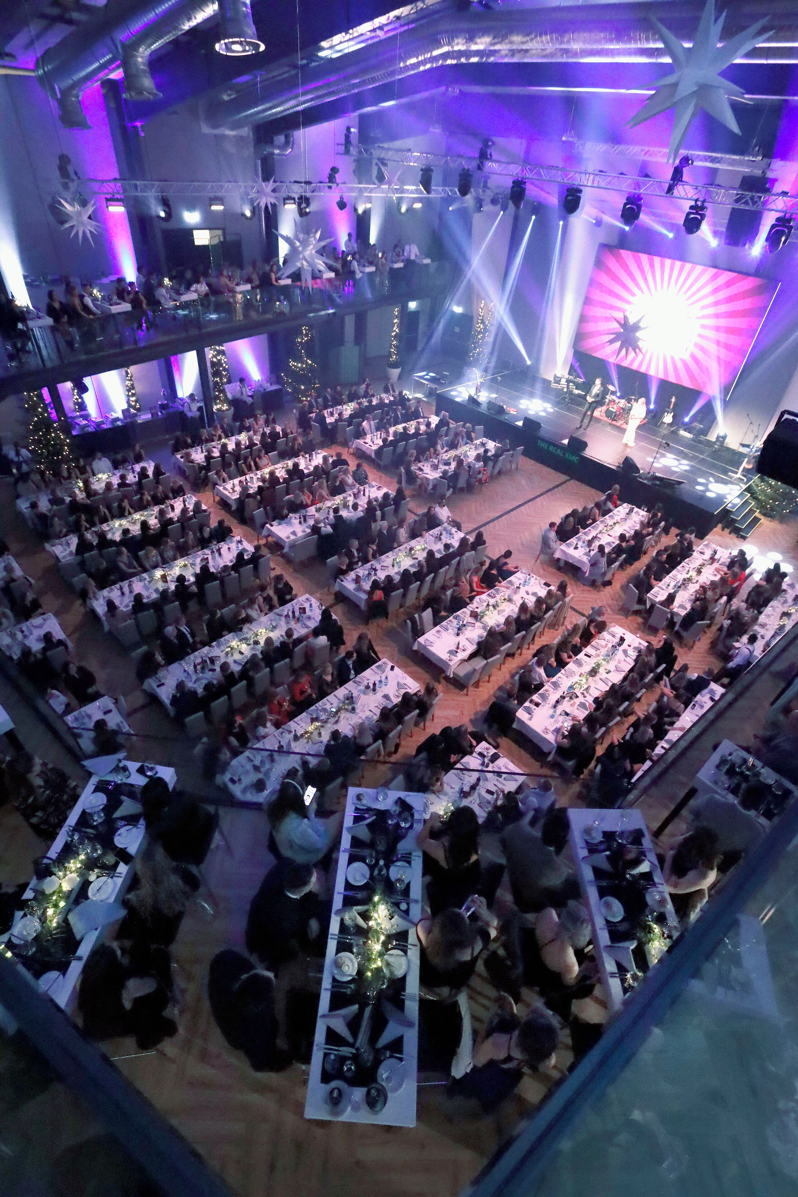 A large indoor event space with multiple tables set up for dining, a stage with performers, and a festive atmosphere with star decorations.