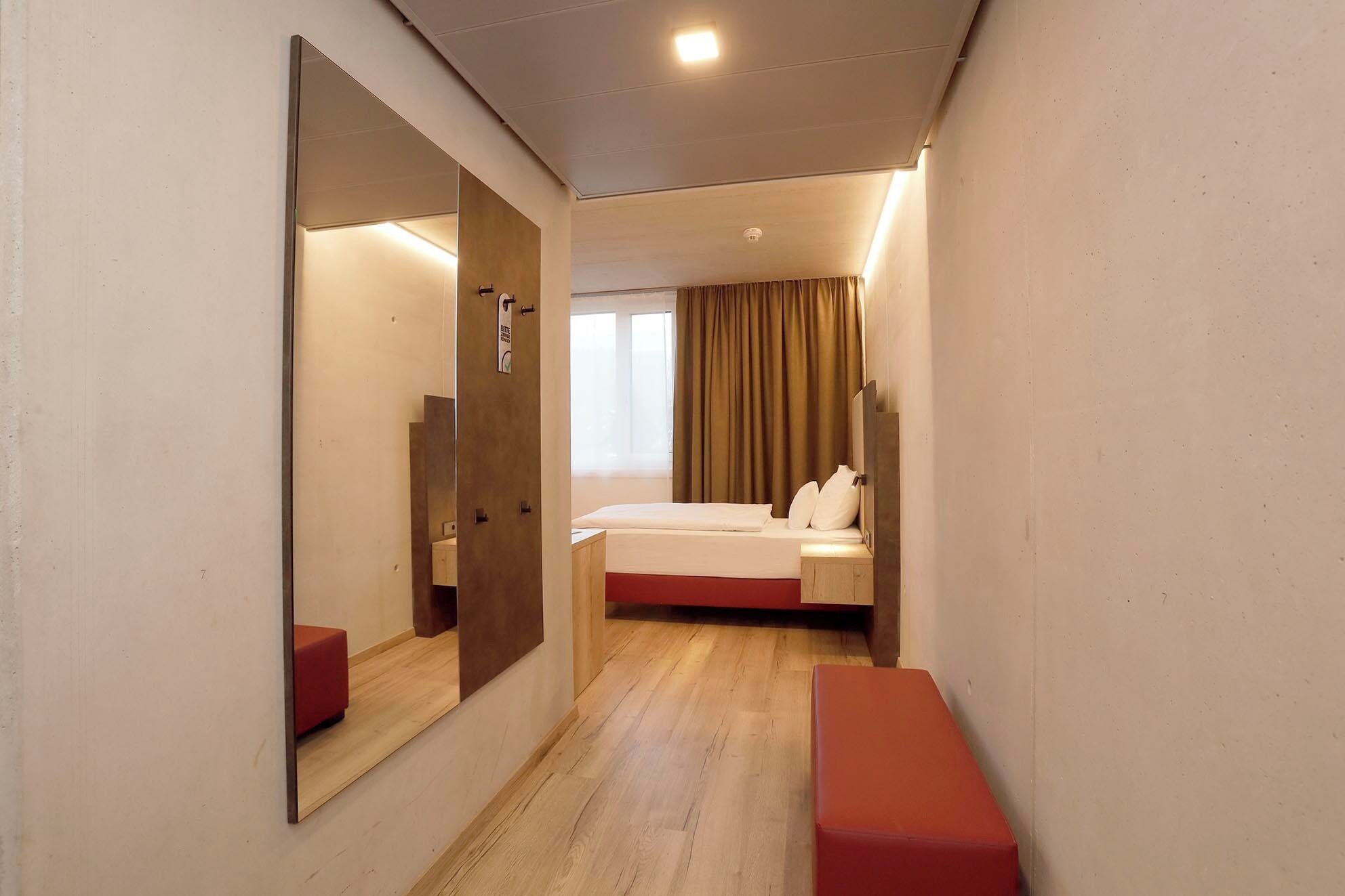 A hotel room with a bed, a bench, a large mirror, and a window with curtains.