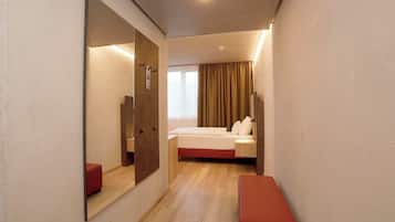 A hotel room with a bed, a bench, a large mirror, and a window with curtains.