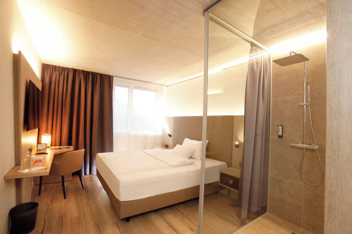 Una habitación de hotel con cama, escritorio, silla y zona de ducha con paredes de vidrio.